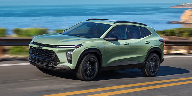 A light green Chevy Trax SUV with a black grille drives along a coastal road with the ocean in the background.